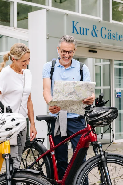 Fahrradtourplan vor Kur & GästeService Ein Mann und eine Frau stehen mit zwei Fahrrädern vor einem Gebäude. Der Mann hält eine Karte, während die Frau lächelt. Die Szene zeigt regionale Gastfreundschaft.