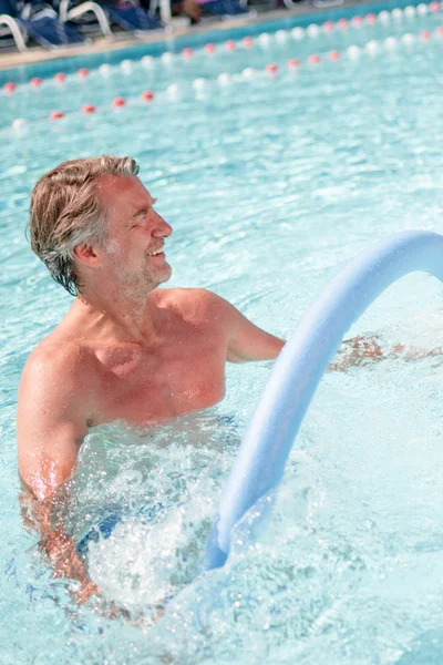 Aktiv im Schwimmbecken Ein Mann und eine Frau stehen im Wasser eines Schwimmbeckens und halten Wasserhanteln. Beide lächeln und scheinen Spaß zu haben. Im Hintergrund sind Liegen und eine Wasseroberfläche sichtbar.