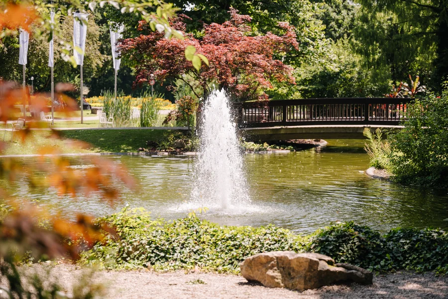 Parkteich mit Brunnen Ein Parkteich mit springendem Brunnen, kleiner Brücke, Bäumen und Blumenbeeten; im Hintergrund stehen weiße Fahnen und Stühle.