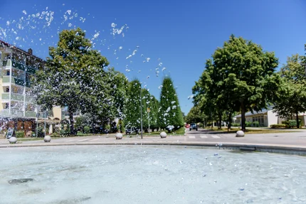 Spritzender Brunnen im Park Ein Brunnen mit spritzendem Wasser, umgeben von Bäumen und Gebäuden, einer klaren blauen Himmel im Hintergrund.
